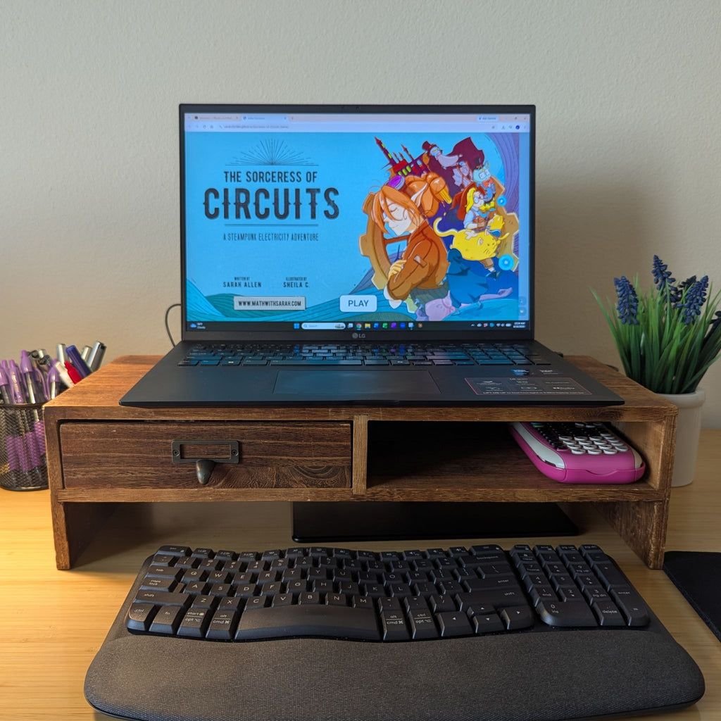A wooden desktop monitor stand holding an open laptop on a desk, with a keyboard and mouse pad in front and a small potted plant to the right. The laptop screen displays colorful artwork and text.