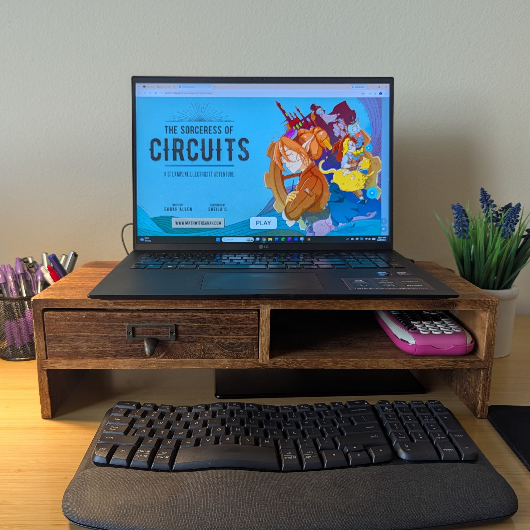 A wooden desktop monitor stand holding an open laptop on a desk, with a keyboard and mouse pad in front and a small potted plant to the right. The laptop screen displays colorful artwork and text.